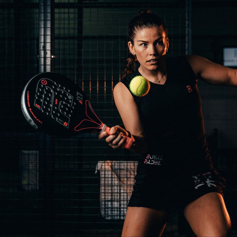 A woman in a dark environment playing padel tennis with a black and red Royal Padel racket